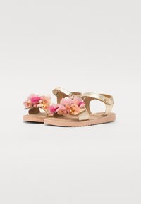 Sandales métalliques dorées avec des embellissements floraux roses, des brides lisses et une semelle texturée couleur tan pour la stabilité et le confort.