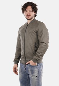 Giacca bomber verde oliva realizzata in morbido suede, caratterizzata da una chiusura frontale con zip, tasca laterale e polsini a costine, abbinata a jeans in denim scoloriti.