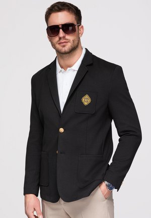 Homme portant un blazer noir avec des boutons dorés et un emblème, une chemise polo blanche, un pantalon beige, des lunettes de soleil et une montre-bracelet, posant devant un fond uni.