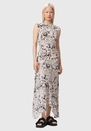 Femme debout portant une robe sans manches, longue jusqu'aux chevilles, à motif abstrait, et des sandales noires à bout ouvert, sur un fond gris uni.