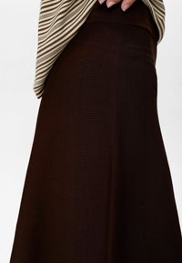 Gros plan sur une personne portant une jupe texturée marron et un haut rayé beige et marron, avec une main reposant près de la taille.