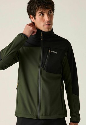 Hombre ajustando el cuello de una chaqueta verde y negra con cremallera, bolsillo en el pecho y logo de Regatta Outdoors sobre fondo claro.