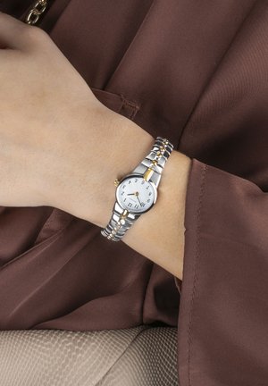 Montre-bracelet en métal argenté avec un cadran blanc, des chiffres noirs, des accents dorés et un bracelet à maillons. Portée sur un poignet contre un fond en tissu brun.