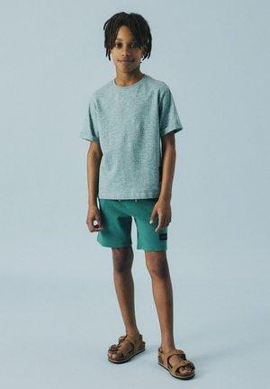 Grå prickig T-shirt, turkosa shorts med logotypmärke och bruna sandaler med remmar. Enkelt mönster, avslappnad passform och mjukt tyg.