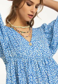 Rochie cu model albastru, decolteu în V, mâneci puff și talie strânsă. Accentuata cu coliere colorate din mărgele stratificate.