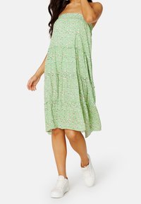 Vihreä kukkainen kes dresses, jossa on kerrostettu muotoilu, pieniä vaaleanpunaisia kukkia, smockattu vartalo ja A-linjainen siluetti, yhdistettynä valkoisiin tennareihin.