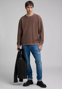 Sudadera marrón con cuello redondo, combinada con vaqueros claros desgastados y botines negros. En una mano sostiene una chaqueta de cuero negra.