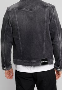 Man med en urblekt svart denimjacka med en Calvin Klein Jeans-etikett på nedre delen av ryggen, kombinerad med en vit skjorta och svarta byxor.