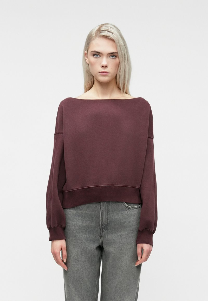 Young person with straight blonde hair wears a loose maroon sweater with wide neckline and faded gray jeans, standing against white background.