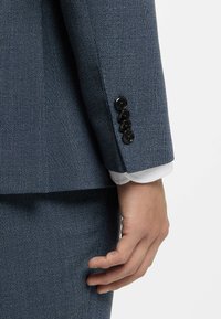 Blå kostymärm med struktur och svarta knappar, med en vit manschett under. Närbild på en hand som vilar mot tyget.