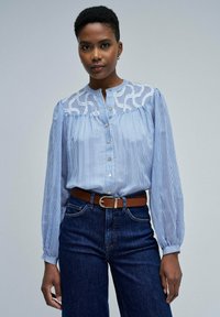 Blau-weiß gestreifte Bluse mit einer Spitzenpasse, langen Ärmeln und Knopfverschluss. Kombiniert mit hochgelegenen, dunklen Denim-Jeans und einem braunen Gürtel.
