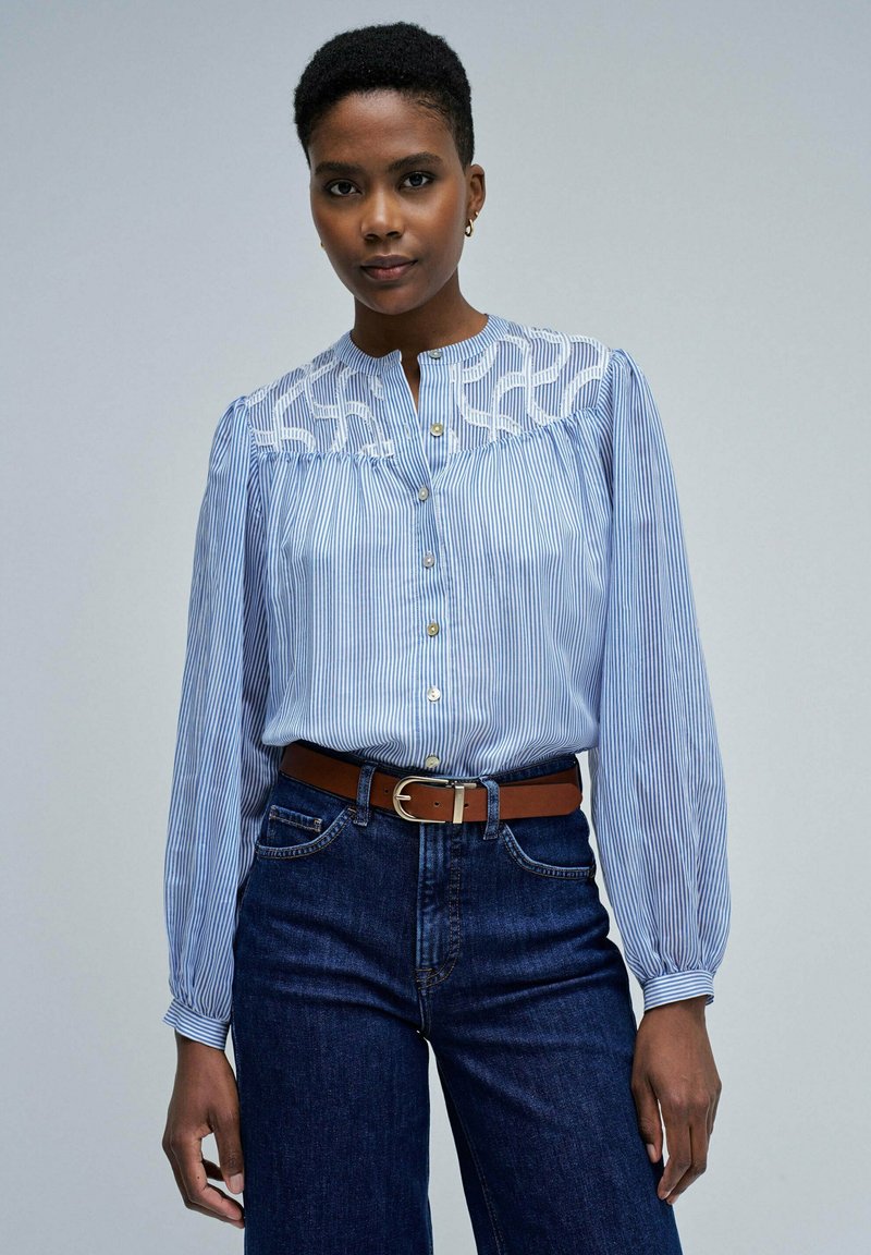 Blau-weiß gestreifte Bluse mit einer Spitzenpasse, langen Ärmeln und Knopfverschluss. Kombiniert mit hochgelegenen, dunklen Denim-Jeans und einem braunen Gürtel.
