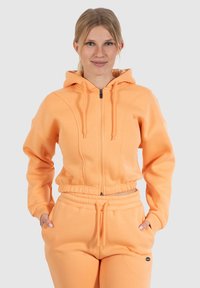 Haut à capuche zippé orange avec une capuche doublée, des poignets côtelés et une taille avec cordon de serrage. Tissu doux avec des poches latérales et des détails de coutures distinctifs.