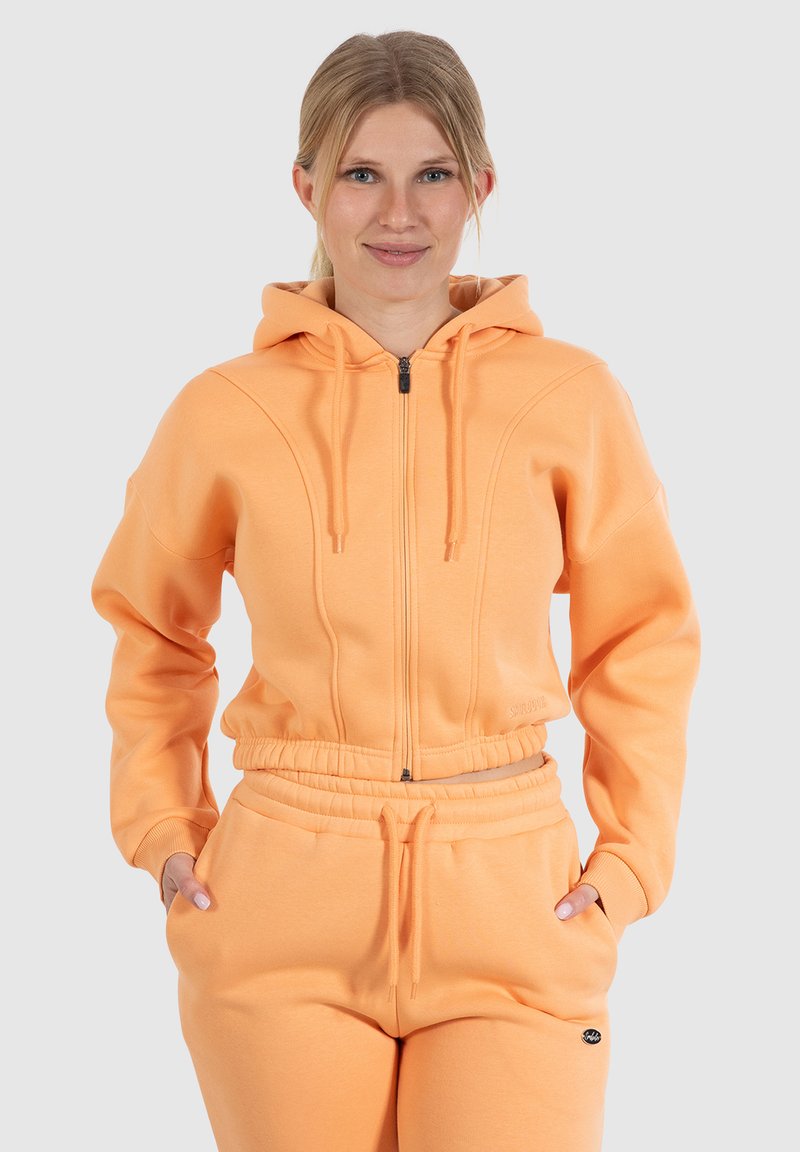 Haut à capuche zippé orange avec une capuche doublée, des poignets côtelés et une taille avec cordon de serrage. Tissu doux avec des poches latérales et des détails de coutures distinctifs.