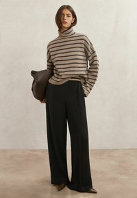 Beżowy sweter z ciemnymi paskami, z golfem. Czarne, szerokie spodnie z wysokim stanem. Modelka trzyma brązową torbę o gładkiej fakturze.