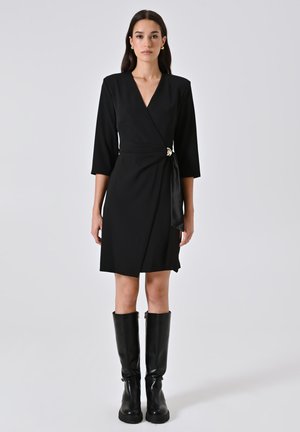 Frau in einem schwarzen knielangen Wickelkleid mit 3/4-Ärmeln und goldener Schnalle, die schwarze knielange Stiefel trägt, vor einem schlichten Hintergrund.