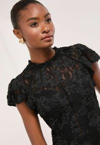 Rochie din dantelă neagră cu aplicații florale, mâneci scurte bufante, guler înalt și un model texturat. Cercei aurii în formă de cerc completează ținuta.