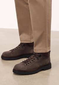 Pantofi din suede maro, cu sireturi, cu un finisaj texturat, talpă din cauciuc negru și accente contrastante de maro închis, asortați cu pantaloni bej.