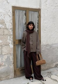 Ruda vilnos mišinio striukė su dviem mygtukais ir šoninėmis kišenėmis, derinama su plačiomis rudomis kelnėmis, rudomis šaliku ir mažos šviesiai rudos odinės rankinės.