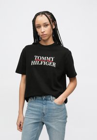 Μαύρο βαμβακερό μπλουζάκι με κοντά μανίκια, με το λογότυπο "TOMMY HILFIGER" σε λευκό και κόκκινο. Σχεδιασμένο με χαλαρή εφαρμογή και στρογγυλή λαιμόκοψη.