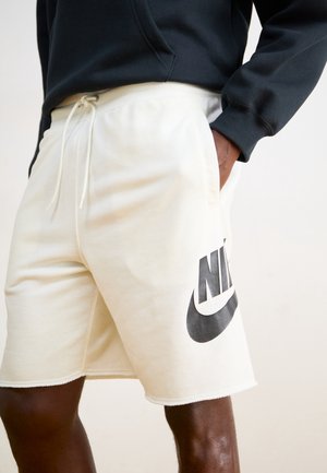 Personne portant un short Nike blanc cassé avec logo noir et cordon, main dans la poche, associé à un haut à manches longues noir.