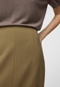 Donna che indossa un top marrone a maniche corte infilato in una gonna verde oliva a vita alta, con una mano appoggiata sul basso schiena.