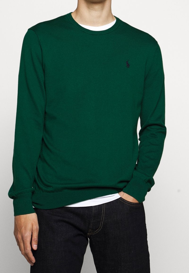 Grön bomullströja i knitstruktur med rund halsringning och ribbade muddarna, med en liten logotyp på vänster bröst. Kombinerad med mörka jeans.