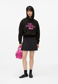 Giovane donna in felpa nera con testo rosa, gonna plissettata nera, calze nere, scarpe nere, mentre tiene una piccola borsa rosa lucida.