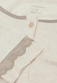 Helles beige Hemd mit Knopfleiste aus weichem Stoff, mit grauem Spitzeborte an den Ärmeln und einem grauen Akzent am Hals. Größe S Etikett sichtbar.