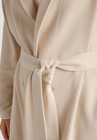Helles beiges Kleid mit gerippter Textur, Schal-Kragen und Bindegürtel in der Taille. Der Stoff wirkt weich und glatt.