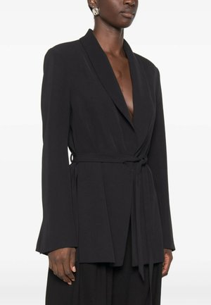 Donna che indossa un blazer nero con cintura, scollo profondo e maniche lunghe, mostrata dalle spalle ai fianchi su sfondo bianco.