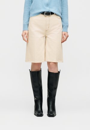 Person trägt beige knielange Shorts, einen hellblauen Pullover, der hineingesteckt ist, einen schwarzen Gürtel und schwarze kniehohe Stiefel und steht vor weißem Hintergrund.