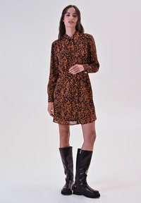 Robe chemise imprimée léopard en noir et orange, à manches longues, avec un devant boutonné et des accents assortis, associée à des bottes noires jusqu'aux genoux.