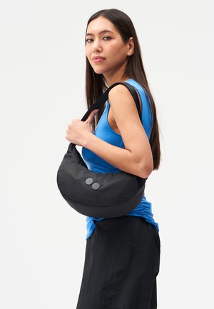 Frau mit langen dunklen Haaren, die ein blaues ärmelloses Oberteil und einen schwarzen Rock trägt, hält eine schwarze, halbmondförmige Umhängetasche mit Knöpfen.