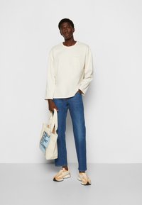 Cremefarvet langærmet skjorte med en lille lomme, blå denim bukser, beige og orange sneakers samt en canvas tote taske med tekstdesign.