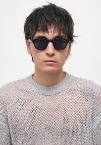 Mustat pyöreät aurinkolasit paksuilla kehyksillä, kerroksellisella tekstuurilla ja kiiltävällä pinnalla. Yhdistettynä vaaleanharmaaseen, löysästi neulottuun villapaitaan.