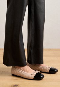 Ballerine beige trapuntate con punta in pelle verniciata nera e dettaglio a fiocco, indossate con pantaloni in pelle neri a gamba larga. Sfondo in parquet.