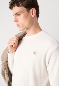 Jeune homme portant un pull blanc texturé avec un petit logo CK, tenant une veste beige sur l'épaule, regardant sur le côté sur un fond uni.