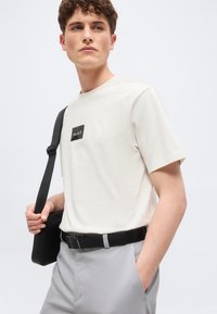 Νεαρός άνδρας με λευκό t-shirt Hugo και ανοιχτό γκρι παντελόνι, κρατώντας μια μαύρη τσάντα ώμου, κοιτάζοντας προς τα πλάγια.