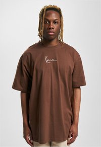 Camiseta de algodón marrón de gran tamaño con mangas cortas, que presenta un logotipo de firma blanco en el pecho. Textura suave con un corte relajado.