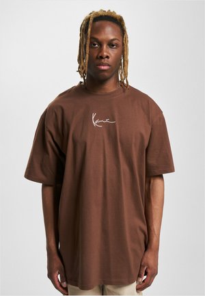 Brown oversized cotton t-shirt with short sleeves, featuring a white signature logo on the chest. Smooth texture with a relaxed fit.