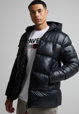 Joven con chaqueta acolchada negra sobre una camiseta blanca con diseño gráfico y fondo gris neutro.