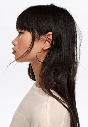 Kobieta o długich czarnych włosach, ubrana w kremową bluzkę, z dużą czarną kolczykiem w kształcie serca, zwrócona w lewo na białym tle.