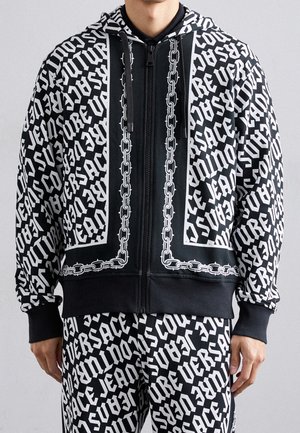 Mężczyzna ubrany w czarno-białą bluzę z kapturem na zamek i pasujące spodnie z powtarzającym się tekstem "VERSACE JEANS COUTURE" i nadrukiem łańcuchów.