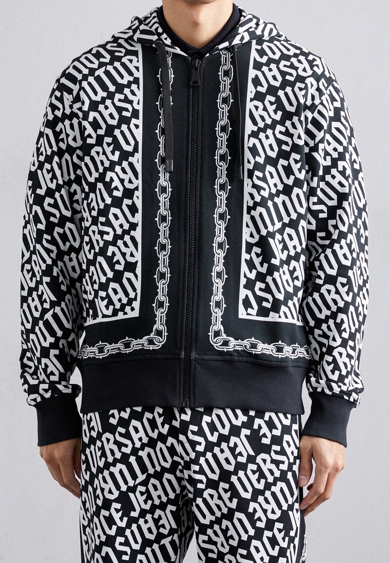 Homme portant un sweat à capuche zippé noir et blanc et un pantalon assorti avec un texte répété "VERSACE JEANS COUTURE" et des accents imprimés à motif chaîne.
