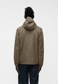 Άνδρας με σκουρόχρωμο δέρμα και πλεξούδες dreadlocks, φορά καφέ μπουφάν με κουκούλα και σκούρα τζιν με κόκκινη κεντητή λεπτομέρεια, γυρισμένος πίσω σε απλό φόντο.