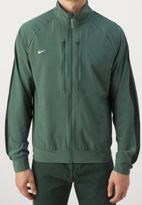 Vihreä urheilu takki, jossa on korkea kaulus, vetoketjukiinnitys edessä, kaksi sivutaskua, mustat sivupaneelit ja pieni valkoinen logo rinnassa.