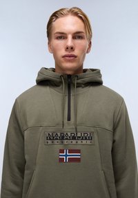 Felpa verde oliva con zip sul collo e cordino, con un grande logo nero del brand e una toppa della bandiera norvegese sul fronte.