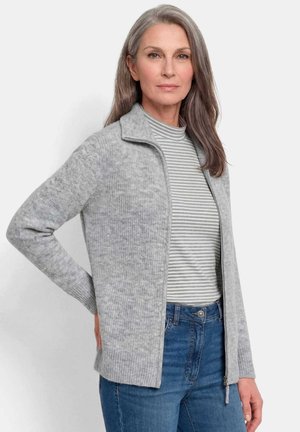 Donna matura con lunghi capelli grigi indossa una maglia grigia con zip sopra una camicia a righe e jeans blu, posando con una mano sul fianco.