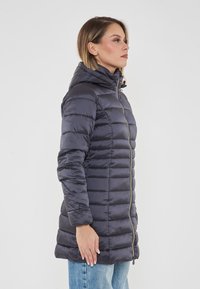 Donna in un cappotto imbottito a maniche lunghe di un grigio scuro con cappuccio, in piedi di lato contro uno sfondo bianco semplice.